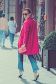 A stylish woman wearing a bright pink coat and sunglasses walks confidently on a street. She holds a matching pink clutch and wears blue jeans with fringed hems, paired with high heels. Her hair is neatly styled, and she exudes confidence as she glances back. The background includes pedestrians and urban architecture, adding to the city vibe.