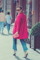 A stylish woman wearing a bright pink coat and sunglasses walks confidently on a street. She holds a matching pink clutch and wears blue jeans with fringed hems, paired with high heels. Her hair is neatly styled, and she exudes confidence as she glances back. The background includes pedestrians and urban architecture, adding to the city vibe.