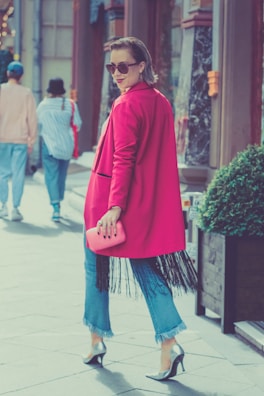 A stylish woman wearing a bright pink coat and sunglasses walks confidently on a street. She holds a matching pink clutch and wears blue jeans with fringed hems, paired with high heels. Her hair is neatly styled, and she exudes confidence as she glances back. The background includes pedestrians and urban architecture, adding to the city vibe.