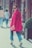 A stylish woman wearing a bright pink coat and sunglasses walks confidently on a street. She holds a matching pink clutch and wears blue jeans with fringed hems, paired with high heels. Her hair is neatly styled, and she exudes confidence as she glances back. The background includes pedestrians and urban architecture, adding to the city vibe.