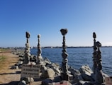 A series of intricately balanced rock towers stand on a rocky shoreline by the water. The towers have several smooth stones stacked vertically. A sign in the foreground requests donations for photos, indicating a possible attraction or artwork.