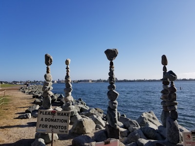 A series of intricately balanced rock towers stand on a rocky shoreline by the water. The towers have several smooth stones stacked vertically. A sign in the foreground requests donations for photos, indicating a possible attraction or artwork.