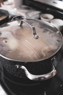 A large metal pot with a speckled surface and stainless steel handles is positioned on a stove. The lid is slightly fogged, indicating that the contents may be steaming or hot. In the background, there are blurred kitchen objects, including two white bowls and another pot.