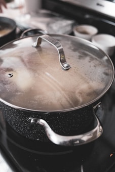 A large metal pot with a speckled surface and stainless steel handles is positioned on a stove. The lid is slightly fogged, indicating that the contents may be steaming or hot. In the background, there are blurred kitchen objects, including two white bowls and another pot.
