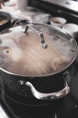 A large metal pot with a speckled surface and stainless steel handles is positioned on a stove. The lid is slightly fogged, indicating that the contents may be steaming or hot. In the background, there are blurred kitchen objects, including two white bowls and another pot.