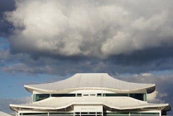 A uniquely designed building with a curved, white roof stands against a dramatic sky filled with large, fluffy clouds.