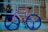 A bicycle with a unique design leans against a light blue railing. The bike features bright blue wheels and a colorful frame with alternating pink and blue stripes. It is positioned on a blue diamond plate ramp next to a brick wall with glass block windows. A potted plant and an open sign are visible in the background through a glass storefront.