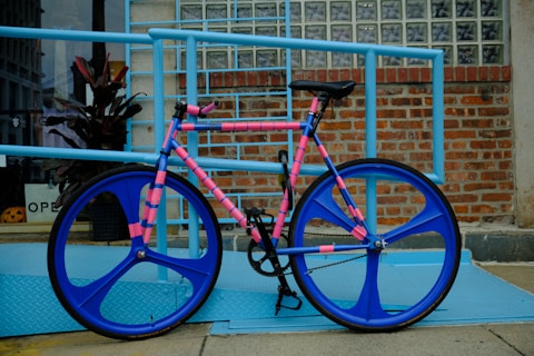 A bicycle with a unique design leans against a light blue railing. The bike features bright blue wheels and a colorful frame with alternating pink and blue stripes. It is positioned on a blue diamond plate ramp next to a brick wall with glass block windows. A potted plant and an open sign are visible in the background through a glass storefront.