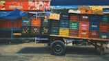 A wooden cart stands on a paved street, loaded with multiple stacks of colorful plastic crates and cardboard boxes. Above this, a vibrant red sign advertises meat products, featuring images of cuts of meat and branding details to the left. The scene has a market or street vendor vibe with hints of blue tarp visible in the background.