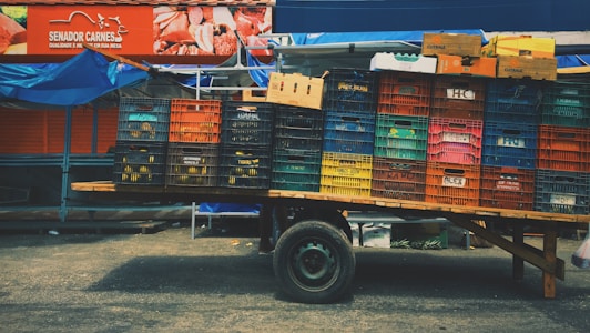 A wooden cart stands on a paved street, loaded with multiple stacks of colorful plastic crates and cardboard boxes. Above this, a vibrant red sign advertises meat products, featuring images of cuts of meat and branding details to the left. The scene has a market or street vendor vibe with hints of blue tarp visible in the background.