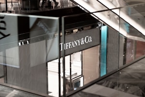 A storefront with the sign 'TIFFANY & CO.' is seen from a higher vantage point. The interior appears to be upscale and modern, featuring glass and metal elements. The store is part of a larger commercial space, indicated by the visible railings and glass panels of a shopping mall.