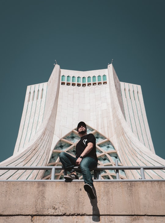 woman in black t-shirt and blue denim jeans sitting on white concrete building during daytime in Azadi Tower Iran