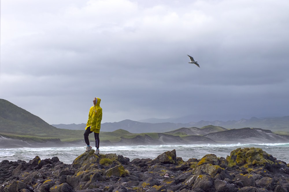 man in yellow hoodie standing on rock near sea during daytime