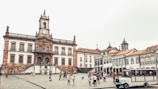 A historic town square featuring a large, ornate colonial building with a clock tower and statues on top. The building has multiple arched windows and a central staircase leading up to the main entrance. Surrounding the square are cobblestone streets and additional colonial-style buildings with red-tiled roofs. People are scattered around, some walking and others gathering in small groups, with a tour bus visible in the foreground.