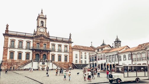 A historic town square featuring a large, ornate colonial building with a clock tower and statues on top. The building has multiple arched windows and a central staircase leading up to the main entrance. Surrounding the square are cobblestone streets and additional colonial-style buildings with red-tiled roofs. People are scattered around, some walking and others gathering in small groups, with a tour bus visible in the foreground.