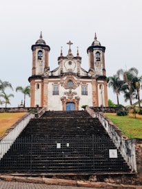 A historic church with a Baroque architectural style features two towers and a detailed facade topped with a cross. The church is approached by a steep staircase flanked by grassy areas and palm trees. The entrance door is a prominent blue color, contrasting with the weathered walls.