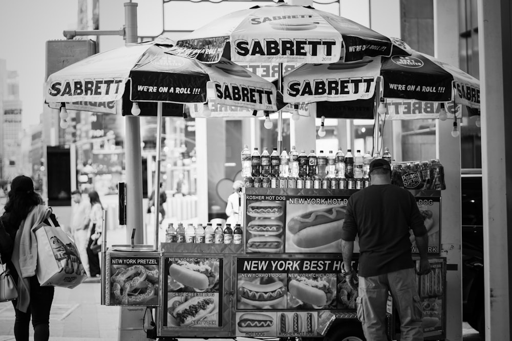 grayscale photo of man in black jacket and pants standing in front of store