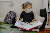 A young child wearing a face mask is reading a picture book while sitting on the floor. The child is seated comfortably against a cushion with a patterned backpack nearby. The setting is indoors, with a small bookshelf to the right containing various books.