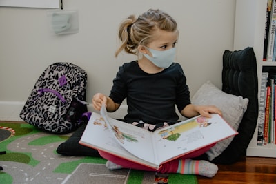A young child wearing a face mask is reading a picture book while sitting on the floor. The child is seated comfortably against a cushion with a patterned backpack nearby. The setting is indoors, with a small bookshelf to the right containing various books.