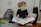 A young child wearing a face mask is reading a picture book while sitting on the floor. The child is seated comfortably against a cushion with a patterned backpack nearby. The setting is indoors, with a small bookshelf to the right containing various books.