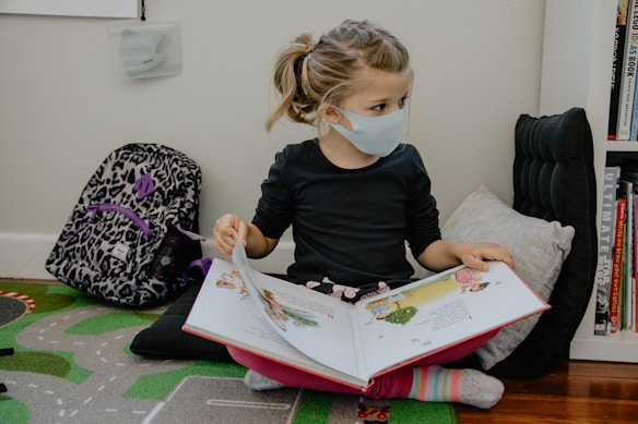 A young child wearing a face mask is reading a picture book while sitting on the floor. The child is seated comfortably against a cushion with a patterned backpack nearby. The setting is indoors, with a small bookshelf to the right containing various books.