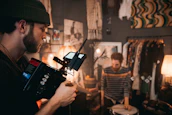 A bearded man wearing a beanie operates a professional video camera in a room filled with various decorative items. In the background, another man is playing on a drum. The setting is cozy, with warm lighting from lamps, and clothing hanging on racks.