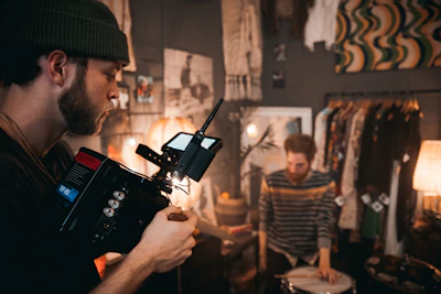 A bearded man wearing a beanie operates a professional video camera in a room filled with various decorative items. In the background, another man is playing on a drum. The setting is cozy, with warm lighting from lamps, and clothing hanging on racks.