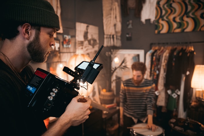 A bearded man wearing a beanie operates a professional video camera in a room filled with various decorative items. In the background, another man is playing on a drum. The setting is cozy, with warm lighting from lamps, and clothing hanging on racks.