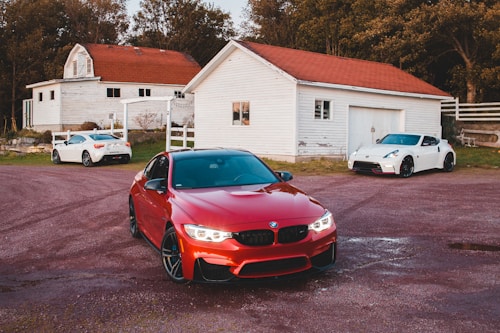 A vibrant red car is prominently displayed in the foreground, parked on a gravel driveway. In the background, there are two white sports cars situated near rustic white barns with red roofs, surrounded by trees, creating a serene countryside setting.