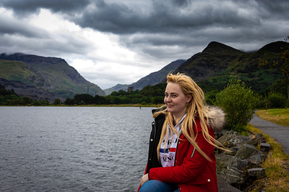 woman in red jacket sitting on rock near body of water during daytime