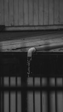 A black and white photograph depicting water flowing out of a pipe mounted on a roof made of corrugated metal. The water is visibly dripping down in a narrow stream, indicating it might be raining. The background shows vertical wooden paneling and part of another structure.