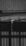 A black and white photograph depicting water flowing out of a pipe mounted on a roof made of corrugated metal. The water is visibly dripping down in a narrow stream, indicating it might be raining. The background shows vertical wooden paneling and part of another structure.