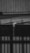 A black and white photograph depicting water flowing out of a pipe mounted on a roof made of corrugated metal. The water is visibly dripping down in a narrow stream, indicating it might be raining. The background shows vertical wooden paneling and part of another structure.