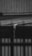 A black and white photograph depicting water flowing out of a pipe mounted on a roof made of corrugated metal. The water is visibly dripping down in a narrow stream, indicating it might be raining. The background shows vertical wooden paneling and part of another structure.
