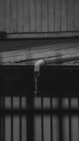 A black and white photograph depicting water flowing out of a pipe mounted on a roof made of corrugated metal. The water is visibly dripping down in a narrow stream, indicating it might be raining. The background shows vertical wooden paneling and part of another structure.