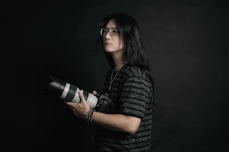 A person with long hair and glasses is holding a professional camera with a large lens. The background is dark, providing a dramatic and focused atmosphere. The subject is wearing a dark, striped shirt and appears to be concentrated or thoughtful.
