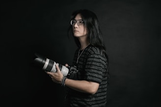 A person with long hair and glasses is holding a professional camera with a large lens. The background is dark, providing a dramatic and focused atmosphere. The subject is wearing a dark, striped shirt and appears to be concentrated or thoughtful.