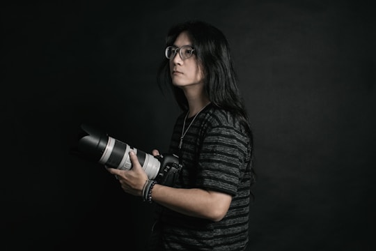 A person with long hair and glasses is holding a professional camera with a large lens. The background is dark, providing a dramatic and focused atmosphere. The subject is wearing a dark, striped shirt and appears to be concentrated or thoughtful.