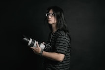 A person with long hair and glasses is holding a professional camera with a large lens. The background is dark, providing a dramatic and focused atmosphere. The subject is wearing a dark, striped shirt and appears to be concentrated or thoughtful.