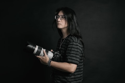 A person with long hair and glasses is holding a professional camera with a large lens. The background is dark, providing a dramatic and focused atmosphere. The subject is wearing a dark, striped shirt and appears to be concentrated or thoughtful.
