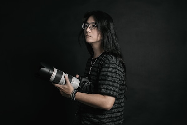 A person with long hair and glasses is holding a professional camera with a large lens. The background is dark, providing a dramatic and focused atmosphere. The subject is wearing a dark, striped shirt and appears to be concentrated or thoughtful.