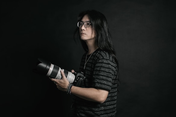 A person with long hair and glasses is holding a professional camera with a large lens. The background is dark, providing a dramatic and focused atmosphere. The subject is wearing a dark, striped shirt and appears to be concentrated or thoughtful.