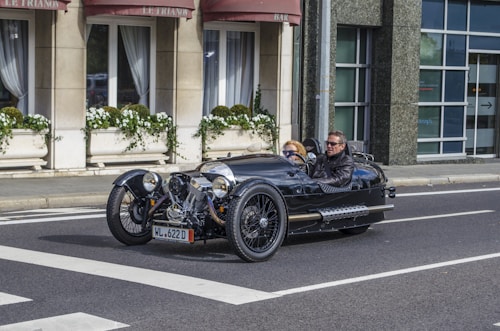 A vintage-style three-wheeled motor car is driving on an urban street. Two individuals are seated in the vehicle. The background features a building with large windows and potted plants, along with awnings that read 'Le Trianon'. The car is glossy black with visible chrome detailing.