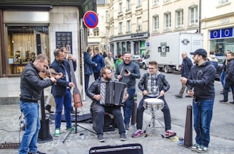 A group of musicians is performing energetically on a city street. The ensemble consists of a violinist, an accordion player, a double bass player, and percussionists. They are surrounded by various people, some watching and others passing by.