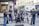 A group of musicians is performing energetically on a city street. The ensemble consists of a violinist, an accordion player, a double bass player, and percussionists. They are surrounded by various people, some watching and others passing by.