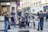 A group of musicians is performing energetically on a city street. The ensemble consists of a violinist, an accordion player, a double bass player, and percussionists. They are surrounded by various people, some watching and others passing by.