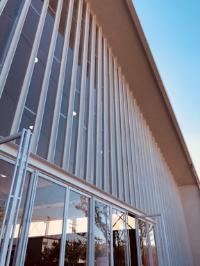 A modern building facade featuring tall vertical white panels, large glass windows, and reflective surfaces. The sky is clear with a light blue color, and some trees and structures are faintly visible in the window reflections.