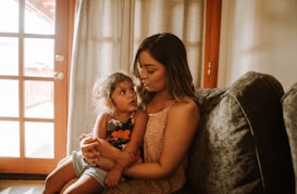 A woman holds a young child on her lap while sitting on a couch in a warmly lit room. They are sharing a tender moment with gentle expressions. The room features a large window with curtains, allowing natural light to enter. The furniture and decor give a cozy atmosphere.