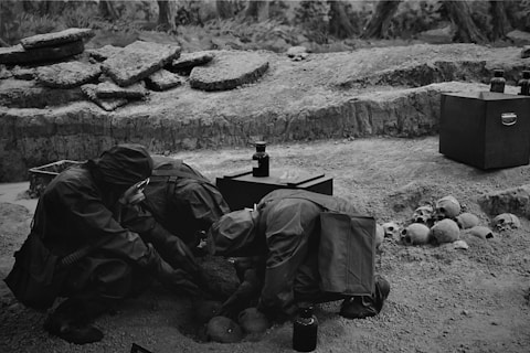 Several individuals in protective suits are examining bones or artifacts in a rough terrain area. The setting appears to be an archaeological or historical excavation site. Various skulls and bones are scattered on the ground along with a few dark-colored containers or tools.
