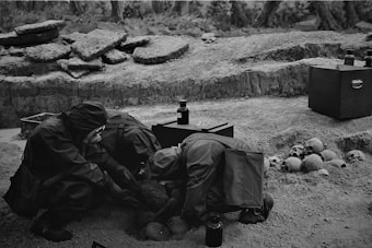Several individuals in protective suits are examining bones or artifacts in a rough terrain area. The setting appears to be an archaeological or historical excavation site. Various skulls and bones are scattered on the ground along with a few dark-colored containers or tools.